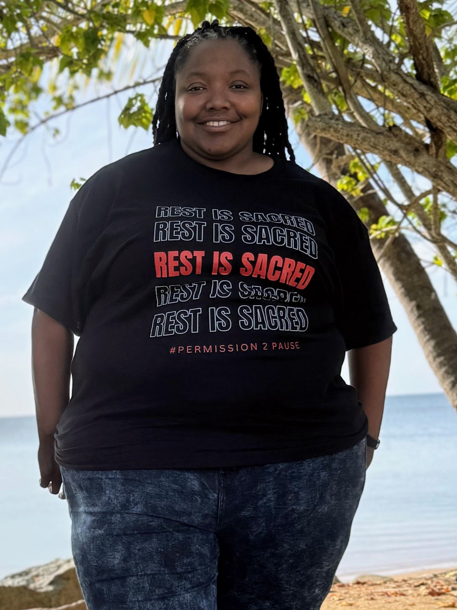 Person wearing a black t-shirt with text standing by trees and water